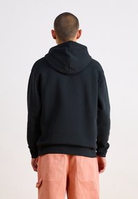 Sudadera negra con un corte relajado, que presenta una capucha grande y ajustable y puños acanalados, combinada con pantalones cargo rosa claro. Diseño sencillo.
