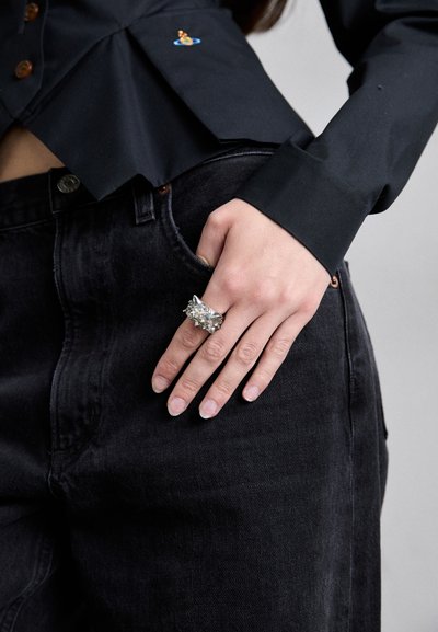 Silver statement ring featuring a textured, multi-faceted design. The wearer has their hand resting on dark denim jeans with a front pocket.