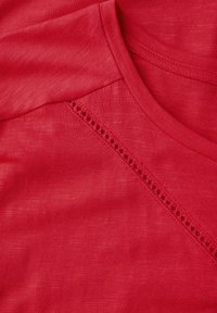 Červené tričko vyrobené z měkké tkaniny, s výstřihem do V a dekorativním háčkovaným detailem na švu ramene. Hladká textura.