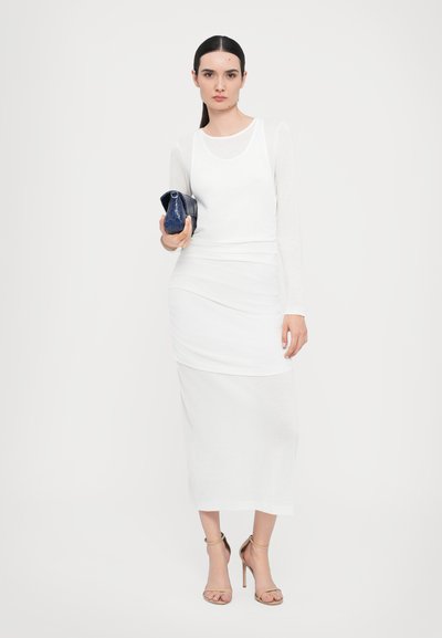 Witte lange mouwen fitted top met een ronde halslijn, geplooide witte rok en beige hoge hakken, terwijl ze een kleine marineblauwe tas met een patroon vasthoudt.