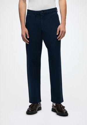 Homme portant un pantalon droit bleu foncé, des chaussettes blanches, des mocassins noirs à pompons et un haut blanc, debout devant un fond uni.