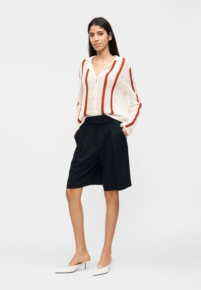 Γυναίκα που στέκεται με τα χέρια στις τσέπες, φορώντας λευκό πλεκτό πουλόβερ με κόκκινες ρίγες, σκούρες μπλε σορτς μέχρι το γόνατο και λευκά mules με τακούνι.