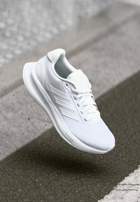 Weißer Sport-Schuh mit einem Mesh-Obermaterial und drei diagonalen Streifen, abgerundeter Zehenpartie, weißen Schnürsenkeln und gepolstertem Sohlen, erhöht über einer Oberfläche.