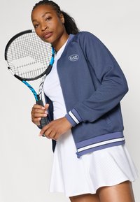 Marineblauwe sportjack met witte en blauwe gestreepte accenten, gecombineerd met een witte rok, vastgehouden met een tennisracket, die een sportieve uitstraling laat zien.