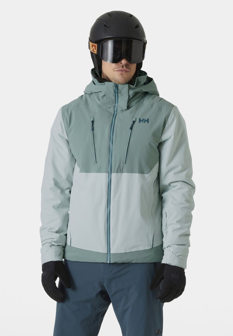 Lichtblauwe skijack met een donkerblauwe bovenkant en twee ritszakken. Ontworpen met een capuchon en gecombineerd met zwarte handschoenen en een skibril.