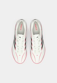 Λευκά ποδοσφαιρικά παπούτσια με κόκκινες λεπτομέρειες και τρεις μαύρες ρίγες. Απαλή υφή, σχεδιασμός γύρου στη μύτη και επίπεδα κορδόνια, εμφανίζονται σε λευκό φόντο.