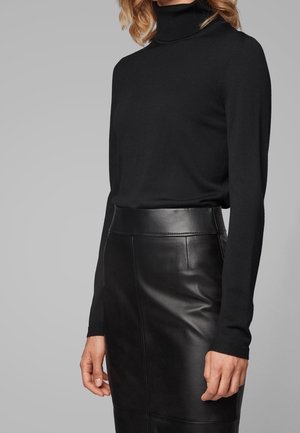 Femme portant un pull à col roulé noir à manches longues, rentré dans une jupe en cuir noire taille haute, debout devant un fond gris.