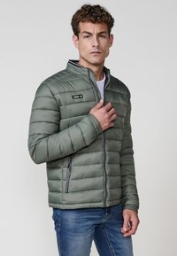 Pūkaina zaļa jaka ar vertikālu quilted dizainu, augstu apkakli un rāvējslēdzēja kabatām. Satur priekšējo rāvējslēdzēju un kontrastējošas apdare detaļas.