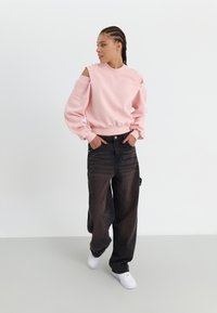 Rosa Sweatshirt mit ausgeschnittenen Ärmeln, passendem Bündchen; kombiniert mit schwarzen weiten Jeans und weißen Sneakern. Glatter Stoff und lockere Passform.