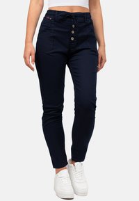 Pantalons bleu marine, fuselés avec taille élastique, fermeture à bouton et poches avant. Portés avec des baskets blanches, mettant en avant un design ajusté.