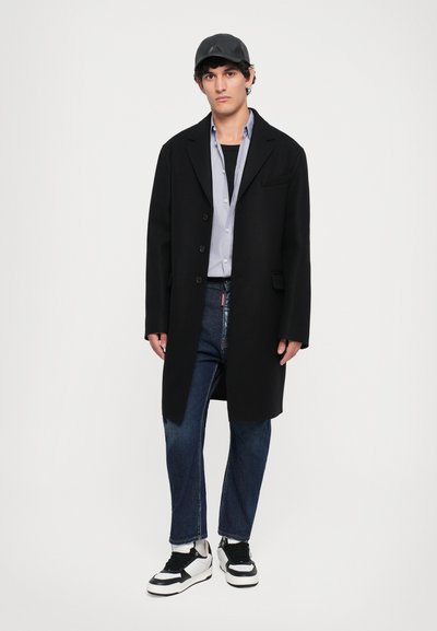 Black coat, light blue shirt, dark jeans, and black-and-white sneakers. Man stands against a plain background. Cap adds detail to outfit.