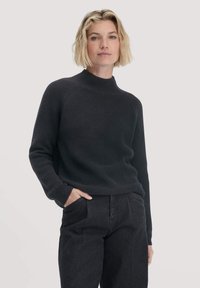 Schwarzer gerippter Strickpullover mit hohem Kragen und langen Ärmeln. Getragen mit dunklen Hosen, die Seitentaschen und einen lockeren Schnitt haben.