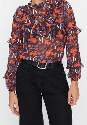 Przezroczysta bluzka z długim rękawem z czerwono-fioletowym kwiatowym wzorem, falbankami na rękawach i piersiach, w połączeniu z czarnymi spodniami z paskiem.