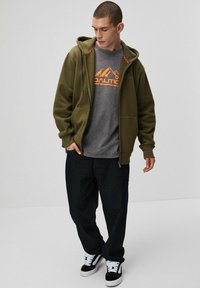 Oliwkowa bluza z zamkiem na całej długości, z wzorzystym sznurkiem, noszona na szarym t-shircie z grafiką, w połączeniu z ciemnymi dżinsami i czarnymi trampkami.