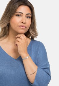 Bracciale d'argento con accenti circolari, indossato su un polso. Maglia azzurro chiaro a maglia con scollatura a V. L'individuo ha capelli mossi lunghi fino alle spalle.