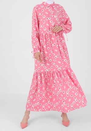 Femme portant une robe maxi fleurie rose à manches longues avec col et jupe à volants, assortie à des chaussures roses à bouts pointus.
