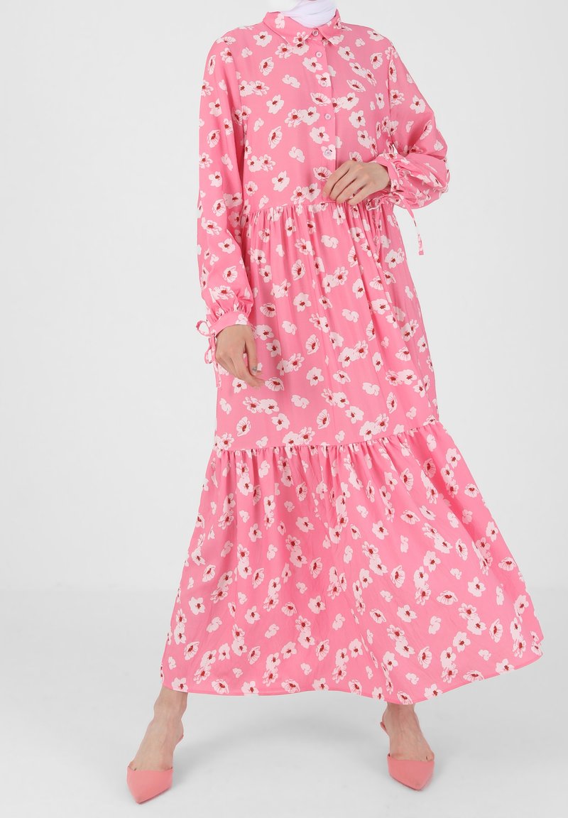 Femme portant une robe maxi fleurie rose à manches longues avec col et jupe à volants, assortie à des chaussures roses à bouts pointus.