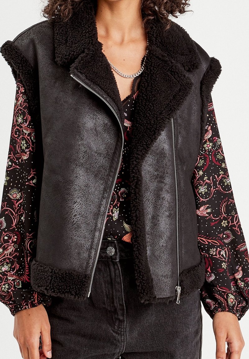 Femme portant un gilet en peau lainée noire sur une blouse fleurie à manches longues, un jean taille haute noir et un collier chaîne argenté.