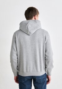 Felpa grigia con un grande cappuccio morbido, orlo e polsini a coste, e una texture liscia. Cuciture visibili aggiungono dettaglio. Indossata con jeans blu.