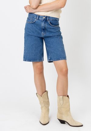 Person trägt blaue Jeans-Shorts mittlerer Länge mit genieteteten Taschen, ein cremefarbenes Stricktop und beige Wildleder-Cowboystiefel, steht mit verschränkten Armen.