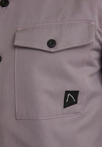 Gros plan d'une poche de chemise mauve avec une fermeture à bouton noir et un petit écusson noir avec un design abstrait blanc.