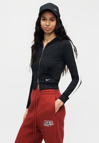Μαύρο κοντό φούτερ με φερμουάρ και λευκές πλαϊνές ρίγες, σε συνδυασμό με κόκκινη φόρμα με το κείμενο "NIKE CLUB" και ελαστικό κορδόνι στη μέση. Μαύρο καπέλο.