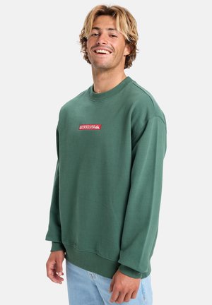 Smiling man with wavy blond hair wears green Quiksilver sweatshirt and light blue jeans, standing against a plain white background.