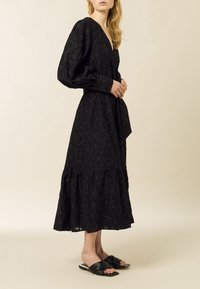 Longue robe noire avec un décolleté en V, des manches longues, un tissu texturé, un ourlet évasé et une ceinture assortie ; assortie à des sandales noires à glissière.