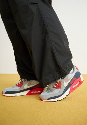 Persona con pantaloni neri e scarpe Nike Air Max grigie, rosse e blu navy in piedi su una superficie gialla.