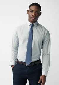 Lichtblauw gestreept overhemd met een button-down kraag, gecombineerd met een blauwe textueltas, een bruine riem en donkerblauwe pantalon.
