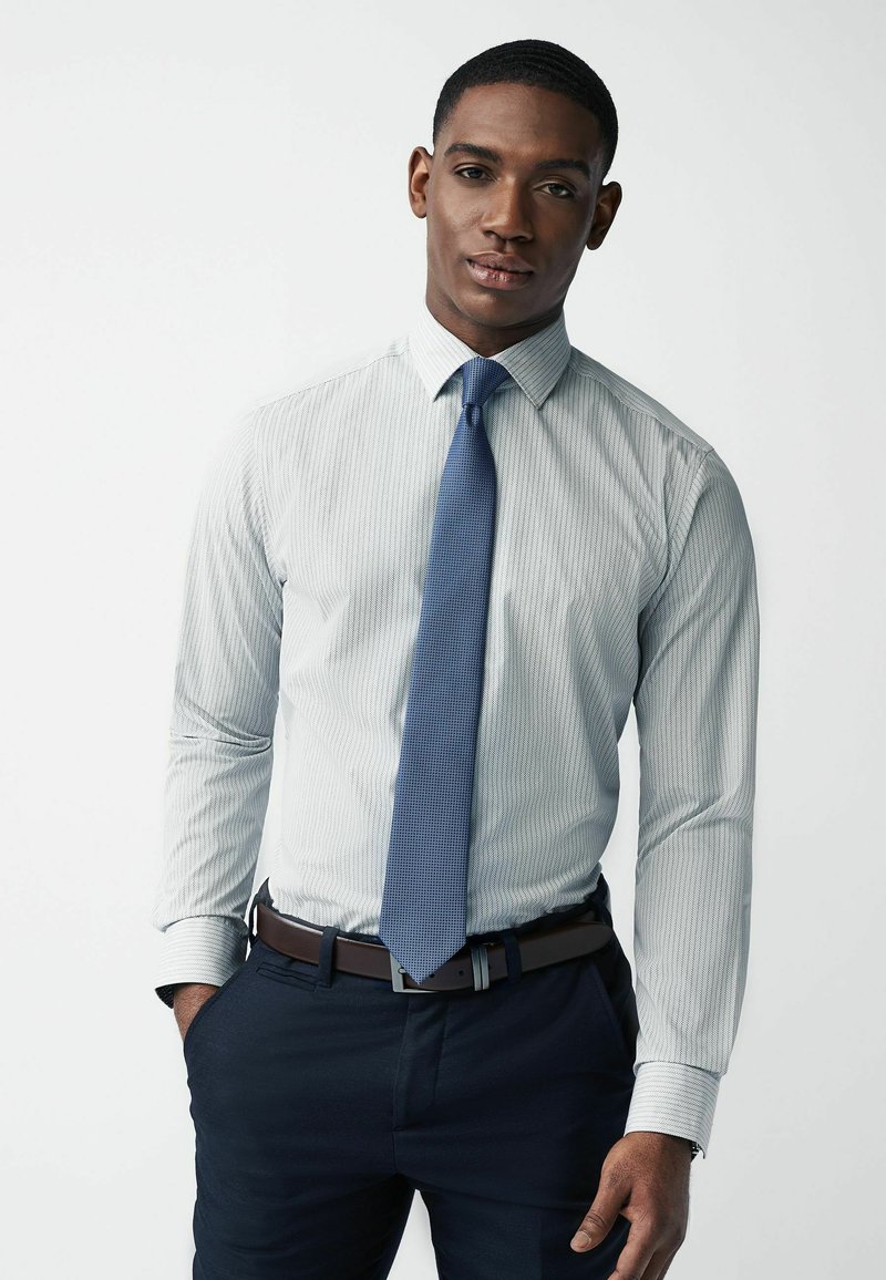 Lichtblauw gestreept overhemd met een button-down kraag, gecombineerd met een blauwe textueltas, een bruine riem en donkerblauwe pantalon.