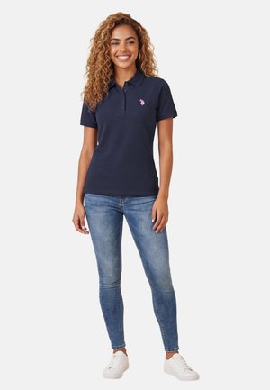 Lächelnde Frau mit lockigem Haar, die ein marineblaues Poloshirt, blaue Jeans und weiße Turnschuhe trägt, vor einem einfarbigen hellen Hintergrund steht.