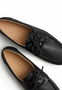 Scarpe in pelle nera con design a stringhe, che presentano un morbido rivestimento interno e sottili dettagli di cucitura. Marca "BALAGAN" impressa all'interno.
