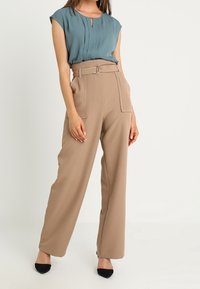 Blouse bleu sarcelle clair à manches courtes avec détails plissés, associée à un pantalon beige taille haute avec ceinture et coupe pattes d'éléphant.