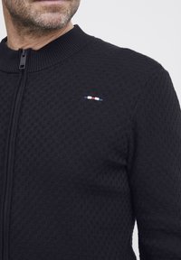 Svart zip-up genser med teksturert mønster, ribbestrikket krage, og en liten tri-farge logo detalj i rødt, hvitt og blått på brystet.