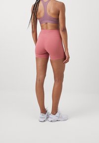 Sujeción deportiva elástica en un tono púrpura suave combinada con shorts rosa de talle alto y ajustados. El material parece suave con un ligero estiramiento. Zapatillas deportivas blancas.
