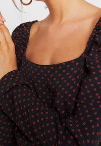 Svart tygblus med röda blommönster, fyrkantig halsringning och puffärmar. Materialet har en mjuk textur med en lätt lyster.