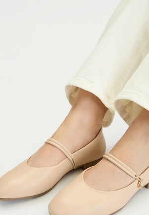 Beżowe buty Mary Jane na niskim obcasie z cienkimi podwójnymi paskami, noszone z jasnokremowymi spodniami na neutralnym tle.