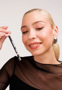 Mustan ja beigen sävyinen meikkikynä, jota henkilö pitää kädessään. Kynässä on tyylikäs muotoilu ja kirkas korkki. Malli pukeutuu läpikuultavaan mustaan toppiin ja suuriin korvakoruihin.