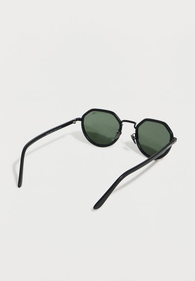Οκταγωνικά γυαλιά ηλίου με μαύρο πλαίσιο και σκουροπράσινους φακούς, εμφανιζόμενα από πλάγια γωνία σε λευκή επιφάνεια.