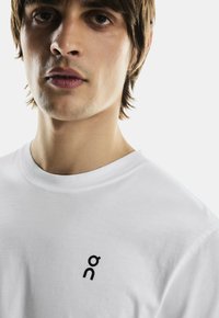 Vit bomulls t-shirt med rund hals, med en liten svart logotyp på vänster sida. Slät textur, figurnära design.