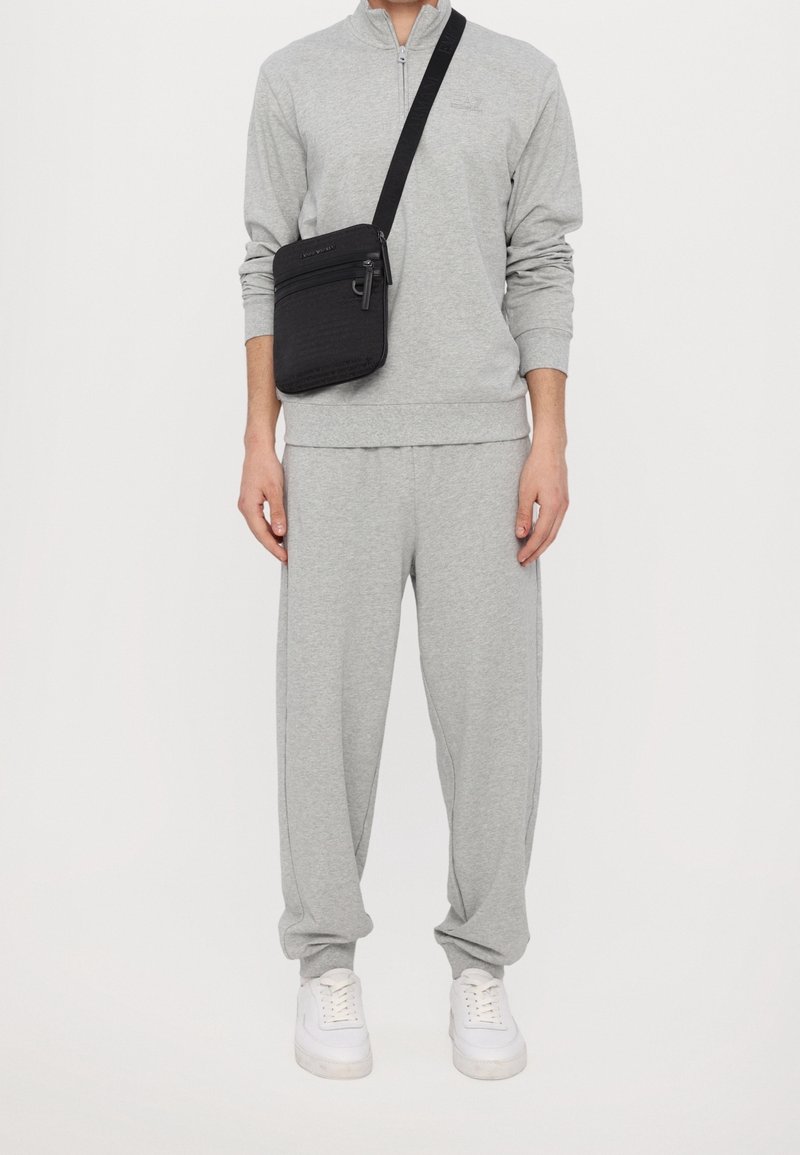 Šedý bavlněný teplákový komplet s dlouhými rukávy a zipem, doplněný o bílé tenisky a černou crossbody tašku se zipy a popruhy.