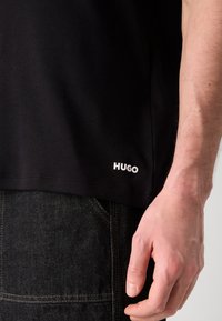 Gros plan d'une personne portant un jean noir et un t-shirt noir avec l'inscription "HUGO" près de l'ourlet, la main droite visible.
