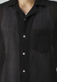 Uomo che indossa una camicia nera a maniche lunghe, in tessuto leggero e con bottoni, con una tasca sul petto sopra una maglietta bianca, mostrato dalla zona del collo fino a metà torace.