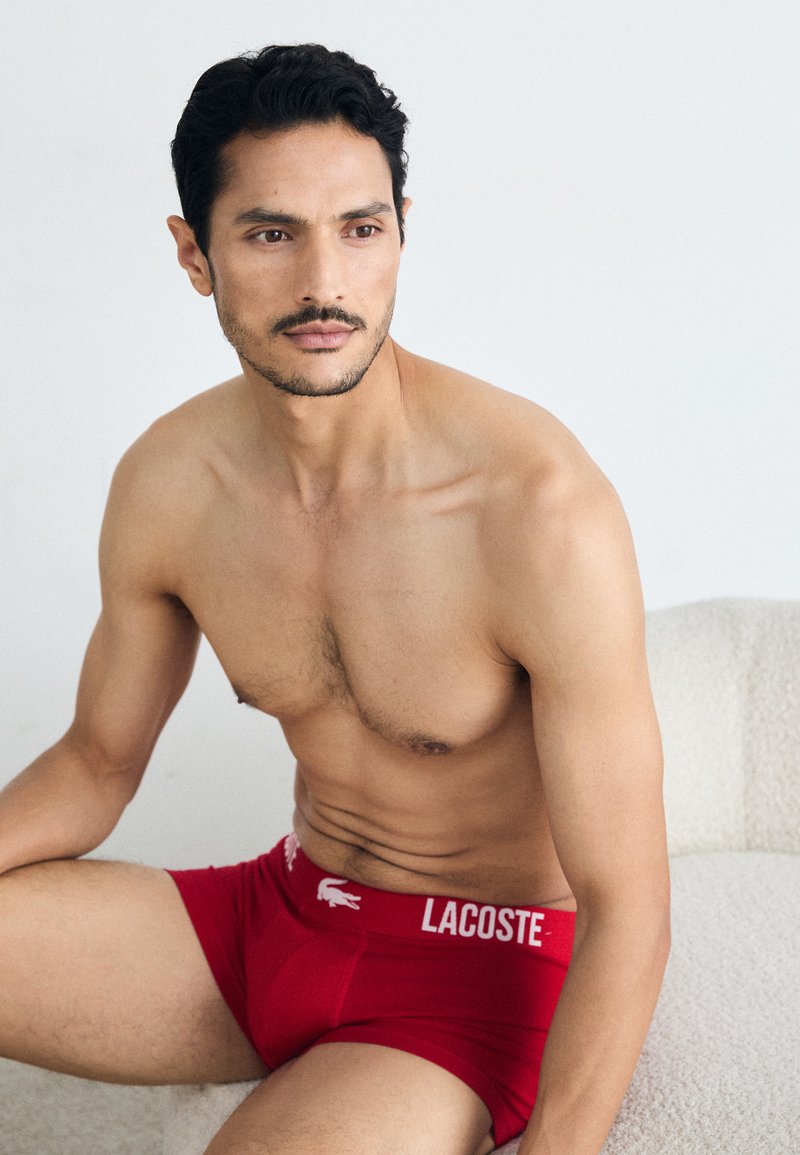 Red Lacoste boxer briefs with white logo at the waistband, sitting on a light textured surface, displaying a smooth garment finish.