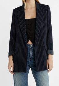 Marineblauw blazer met krijtstreep, een getailleerde pasvorm, revers met inkeping en omgeslagen mouwen, gecombineerd met een cropped zwart topje en lichtblauwe spijkerbroek.