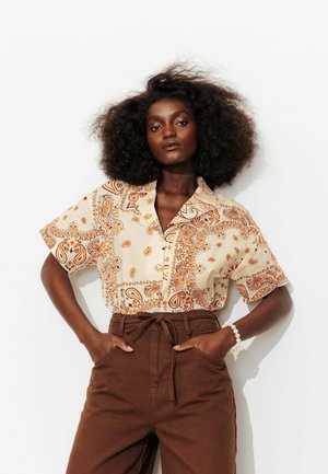 Femme aux cheveux bouclés et volumineux porte une chemise beige à manches courtes à motif cachemire rentrée dans un pantalon marron taille haute, mains dans les poches, bracelet blanc.