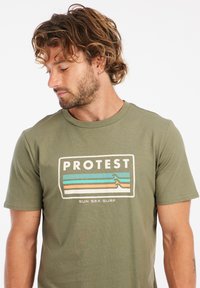 Alyvuogių žalios spalvos medvilnės marškiniai, su stačiakampiu logotipu, kuriame užrašyta "PROTEST" ir banguotomis linijomis mėlyna, oranžine ir ruda spalvomis. Trumpomis rankovėmis.