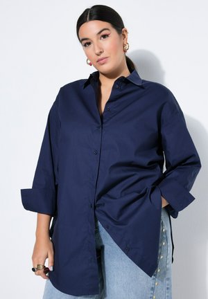 Vrouw met donker haar die gouden oorringen draagt, een oversized marineblauw overhemd met knopen en lichtblauwe jeans met gouden knopen.