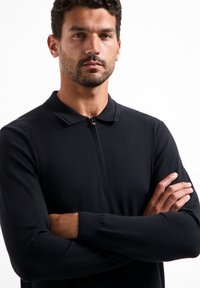 Zwarte langwerpige polo met lange mouwen en een halve rits-kragen, gemaakt van een gladde, gestructureerde stof. Heeft een slim fit en geribbelde manchetten.